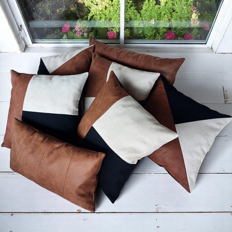May include: A collection of decorative throw pillows in various shapes and sizes. The pillows feature geometric designs with brown, black, and cream-colored panels. Some pillows have a solid brown leather-like texture. The pillows are arranged on a white wooden surface.