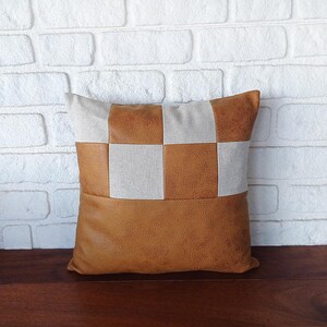 May include: A brown and beige checkered throw pillow with a faux leather bottom. The pillow is made of a soft, textured fabric.