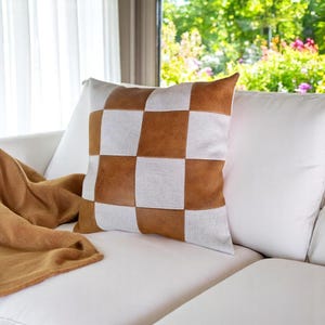 May include: A white and brown checkered throw pillow with a brown leather-like texture. The pillow is on a white couch with a brown throw blanket.