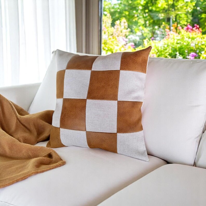 May include: A white and brown checkered throw pillow with a brown leather-like texture. The pillow is on a white couch with a brown throw blanket.