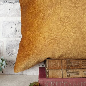 May include: A mustard yellow throw pillow with a textured surface sits on top of a stack of three vintage books. The books are brown and have gold lettering on the spines.