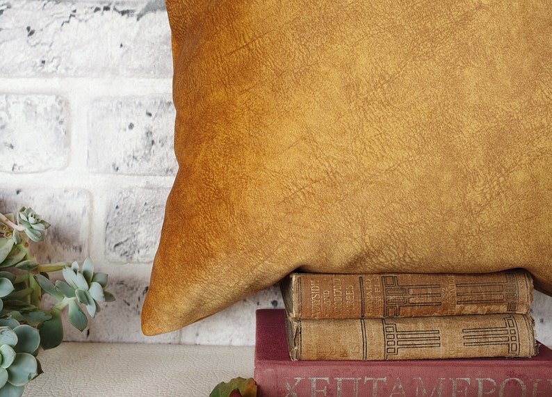 May include: A mustard yellow throw pillow with a textured surface sits on top of a stack of three vintage books. The books are brown and have gold lettering on the spines.