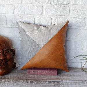 May include: A decorative pillow with a geometric design. One side is made of a light brown faux leather and the other side is a light gray linen fabric. The pillow is sitting on a wooden surface with a book in front of it.
