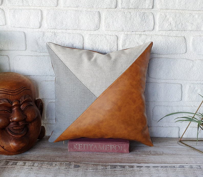 May include: A decorative pillow with a geometric design. One side is made of a light brown faux leather and the other side is a light gray linen fabric. The pillow is sitting on a wooden surface with a book in front of it.