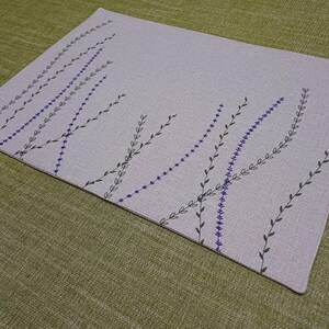 May include: A light gray linen placemat with a pattern of black and purple embroidered sprigs.