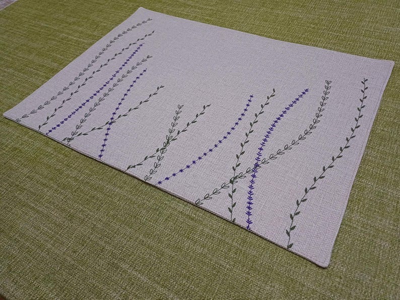 May include: A light gray linen placemat with a pattern of black and purple embroidered sprigs.
