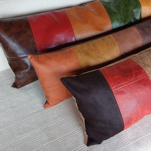 May include: Three rectangular throw cushions featuring a colour-blocked design with panels of faux leather in shades of dark brown, red, orange, yellow, green, and tan. Each cushion has a textured surface and is arranged in a staggered, overlapping row.