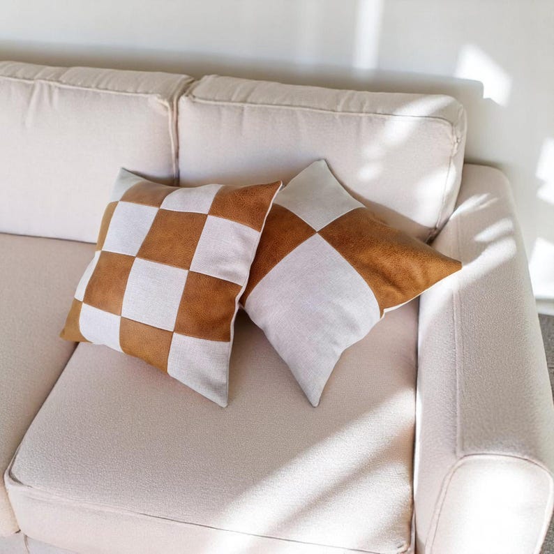 May include: Two decorative throw pillows with a checkered pattern. The pillows are made of a light brown faux leather and a white fabric. The pillows are on a white couch.