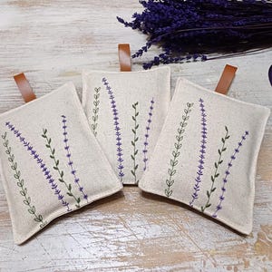 May include: Three white linen sachets with lavender embroidery. The sachets have brown leather ties and are filled with dried lavender.
