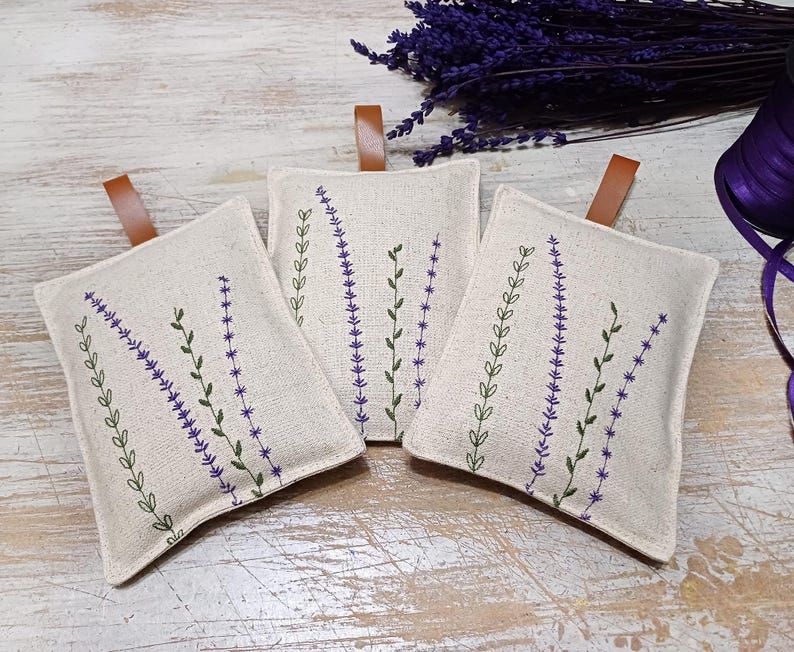 May include: Three white linen sachets with lavender embroidery. The sachets have brown leather ties and are filled with dried lavender.