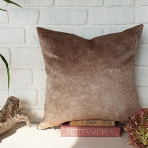 May include: A brown suede pillow with a textured surface. The pillow is sitting on a stack of books with the text "HEPTAMERON" visible.