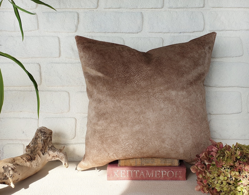 May include: A brown suede pillow with a textured surface. The pillow is sitting on a stack of books with the text "HEPTAMERON" visible.