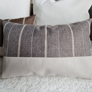 May include: A striped throw pillow with a beige and brown color scheme. The pillow has a white stripe pattern and a beige faux leather bottom.