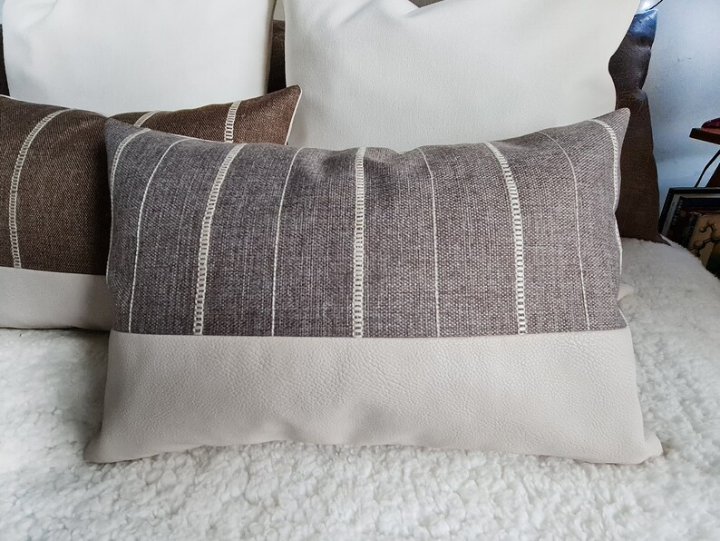 May include: A striped throw pillow with a beige and brown color scheme. The pillow has a white stripe pattern and a beige faux leather bottom.
