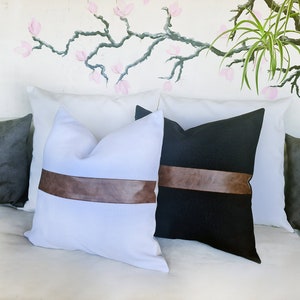Puede incluir: Dos almohadas decorativas con detalles de cuero marr&oacute;n. Una almohada es blanca con una banda de cuero marr&oacute;n, y la otra es negra con una banda de cuero marr&oacute;n.