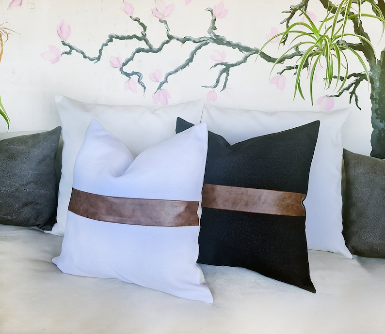 Puede incluir: Dos almohadas decorativas con detalles de cuero marr&oacute;n. Una almohada es blanca con una banda de cuero marr&oacute;n, y la otra es negra con una banda de cuero marr&oacute;n.