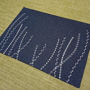 May include: A dark blue placemat with a white embroidered design of stylised leaves and branches.