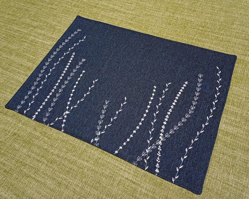 May include: A dark blue placemat with a white embroidered design of stylised leaves and branches.