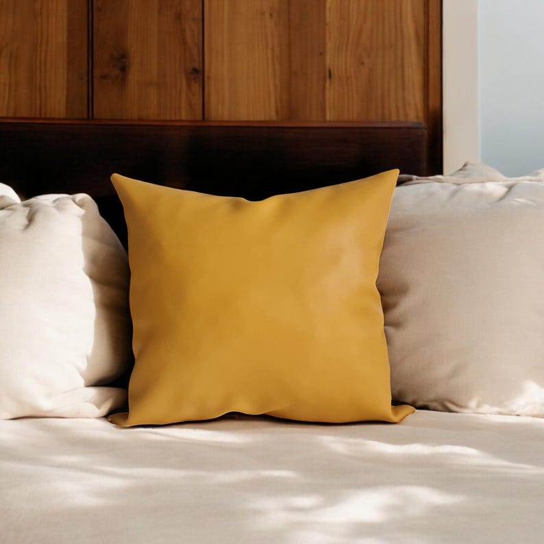 May include: A mustard yellow throw pillow with a smooth, leather-like texture sits on a bed with white bedding.