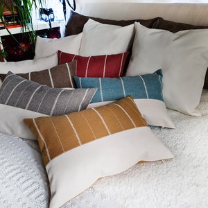 May include: A collection of decorative throw pillows in various colors and patterns, including brown, gray, blue, and red stripes, and a solid white pillow. The pillows are arranged on a white fluffy bedspread.