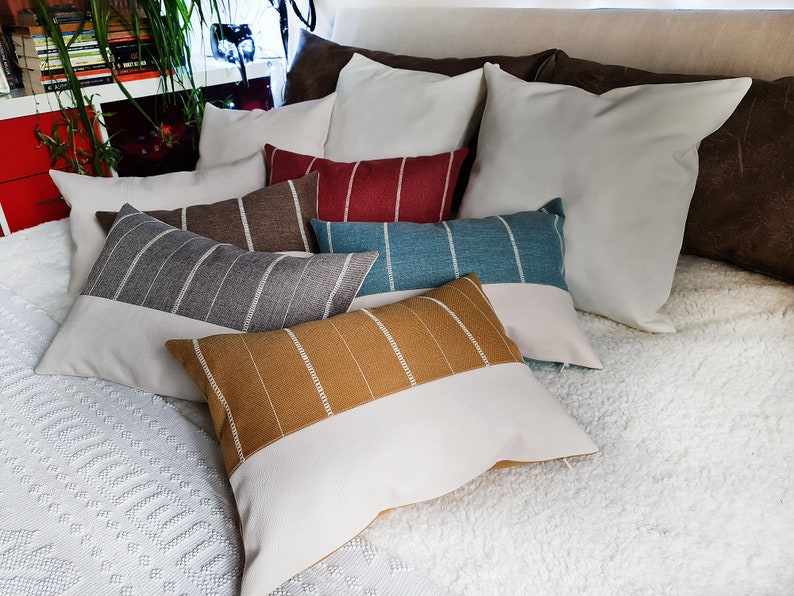 May include: A collection of decorative throw pillows in various colors and patterns, including brown, gray, blue, and red stripes, and a solid white pillow. The pillows are arranged on a white fluffy bedspread.