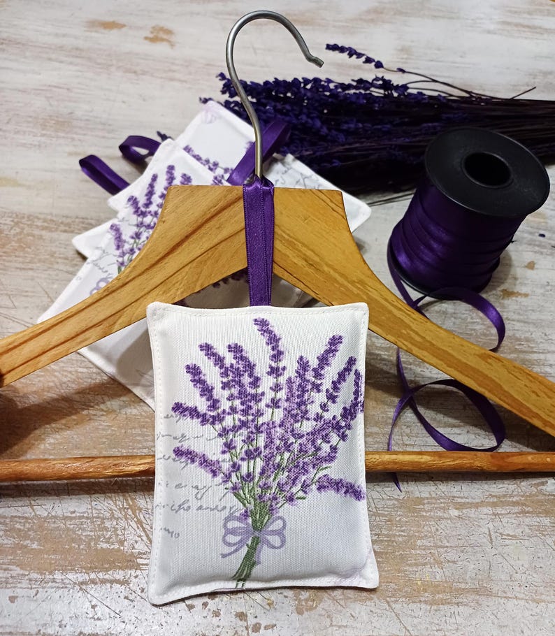 Op de afbeelding: Een wit stoffen lavendelzakje met een paars lavendelboeketontwerp dat aan een houten hanger hangt met een paars lint.