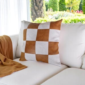 May include: A brown and white checkered throw pillow with a faux leather design. The pillow is on a white couch with a brown throw blanket.