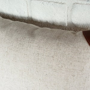 May include: A white linen pillow with brown leather accents. The pillow has a simple, elegant design and is perfect for adding a touch of sophistication to any room.