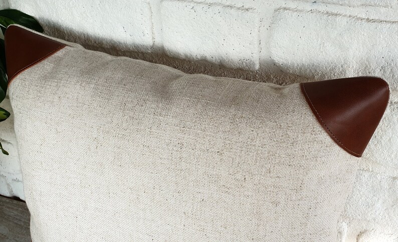 May include: A white linen pillow with brown leather accents. The pillow has a simple, elegant design and is perfect for adding a touch of sophistication to any room.