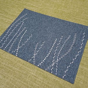 May include: A blue placemat with a white embroidered design of stylised leaves and branches.