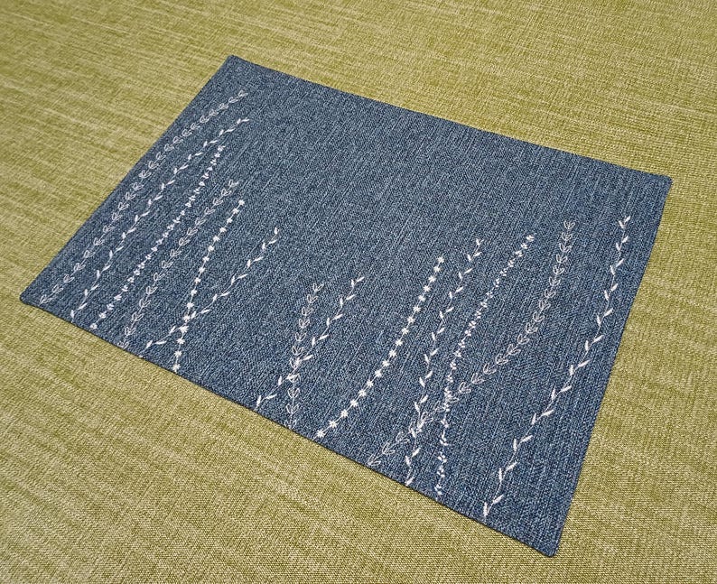 May include: A blue placemat with a white embroidered design of stylised leaves and branches.