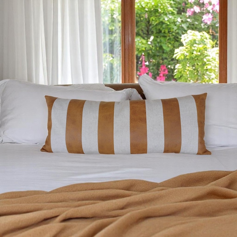May include: A white bed with a brown and white striped lumbar pillow and a brown blanket. The bed is in front of a window with white curtains and a view of green trees.