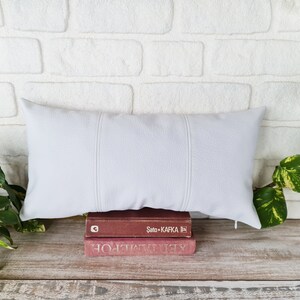 May include: A white rectangular pillow with three stitched seams down the center, resting on top of two books. The top book has the text "Sato - Kafka" printed on the spine. The books are on a light brown wooden surface.