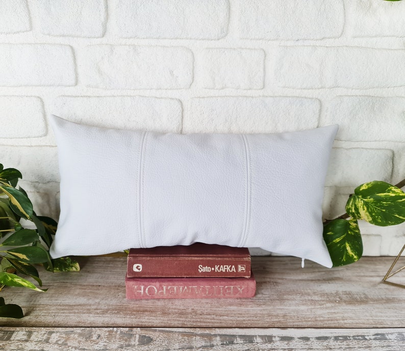 May include: A white rectangular pillow with three stitched seams down the center, resting on top of two books. The top book has the text "Sato - Kafka" printed on the spine. The books are on a light brown wooden surface.
