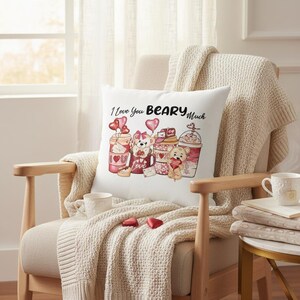 May include: A white square pillow with the phrase "I Love You Beary Much" and illustrations of teddy bears and coffee cups. The pillow is on a beige chair with a cream-colored blanket. Two heart-shaped candies are on the chair.