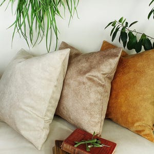 May include: Three decorative throw pillows in various shades of beige and brown. The pillows are made of a soft, textured fabric and are arranged on a white surface.