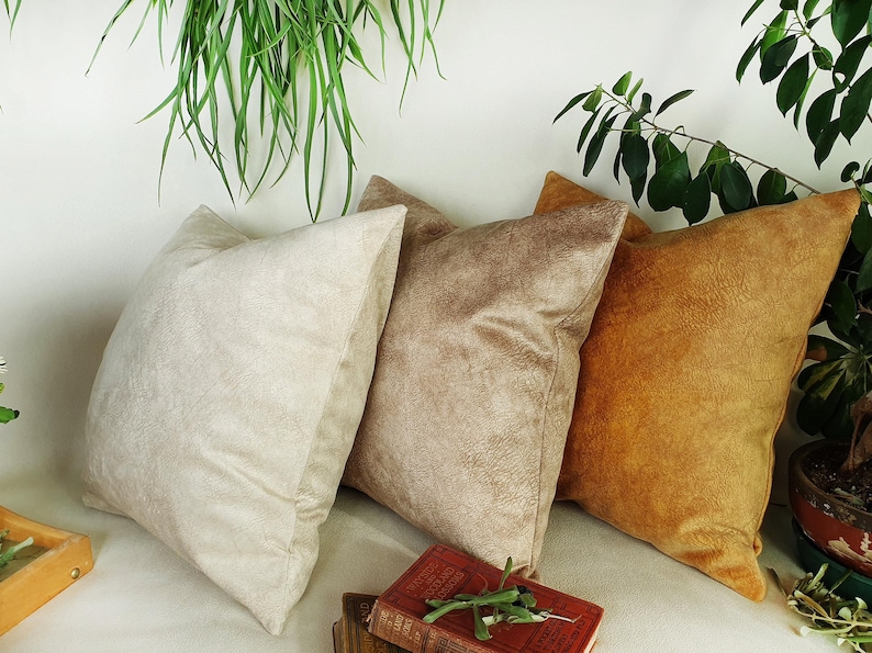 May include: Three decorative throw pillows in various shades of beige and brown. The pillows are made of a soft, textured fabric and are arranged on a white surface.