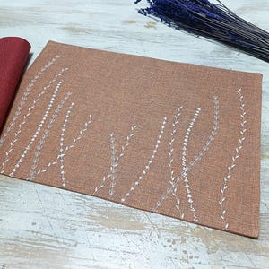 May include: A brown placemat with a white embroidered design of stylized grass or plants. The placemat is made of a textured fabric.