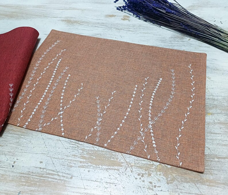 May include: A brown placemat with a white embroidered design of stylized grass or plants. The placemat is made of a textured fabric.
