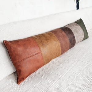 May include: A long rectangular bolster pillow featuring a patchwork design of faux leather panels in various shades of brown, tan, rust, and dark green. The surface has a textured, weathered appearance with visible stitched seams between each color block.