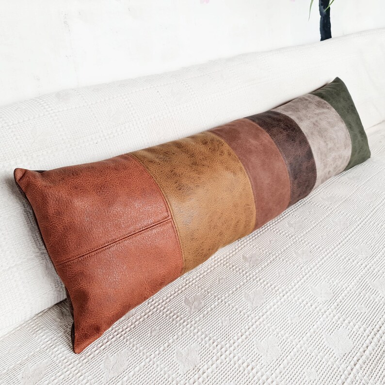 May include: A long rectangular bolster pillow featuring a patchwork design of faux leather panels in various shades of brown, tan, rust, and dark green. The surface has a textured, weathered appearance with visible stitched seams between each color block.