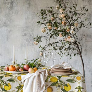 May include: A dining table set for a meal, featuring a tablecloth with a lemon and leaf pattern. The table is set with plates, glasses, and a fruit arrangement. Two white candles stand on the table, with a floral arrangement in the background.