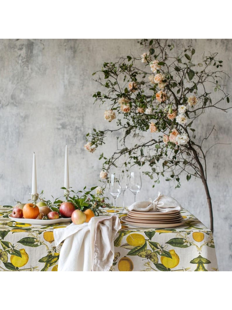 May include: A dining table set for a meal, featuring a tablecloth with a lemon and leaf pattern. The table is set with plates, glasses, and a fruit arrangement. Two white candles stand on the table, with a floral arrangement in the background.