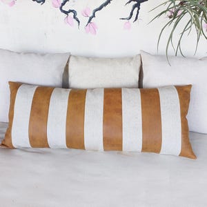 May include: A long, rectangular throw pillow with brown and white stripes. The pillow is made of a soft, textured fabric and has a subtle sheen. The pillow is lying on a white bedspread.