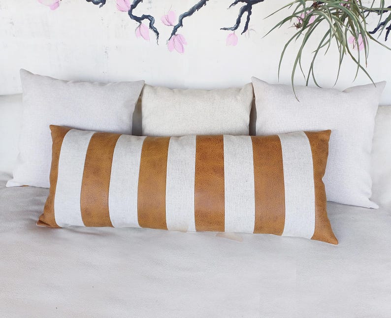 May include: A long, rectangular throw pillow with brown and white stripes. The pillow is made of a soft, textured fabric and has a subtle sheen. The pillow is lying on a white bedspread.
