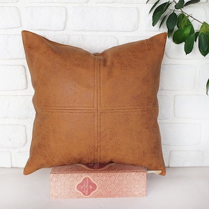 May include: A brown faux leather pillow with a square stitched design. The pillow is sitting on a white surface with a book in front of it.