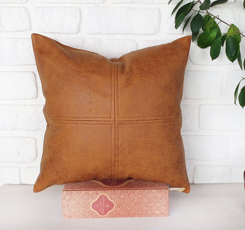May include: A brown faux leather pillow with a square stitched design. The pillow is sitting on a white surface with a book in front of it.
