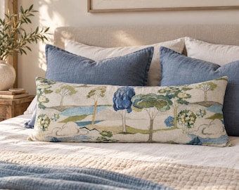 May include: A rectangular lumbar pillow featuring a printed landscape scene with green and blue trees, rolling hills, and white clouds on a light beige fabric. The pillow is positioned in front of two solid blue square pillows on a bed.