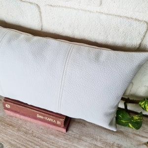 May include: White faux leather lumbar pillow with a subtle textured surface and stitched seams. The pillow is resting on a wooden surface with a book titled "Sato + Kafka" visible.