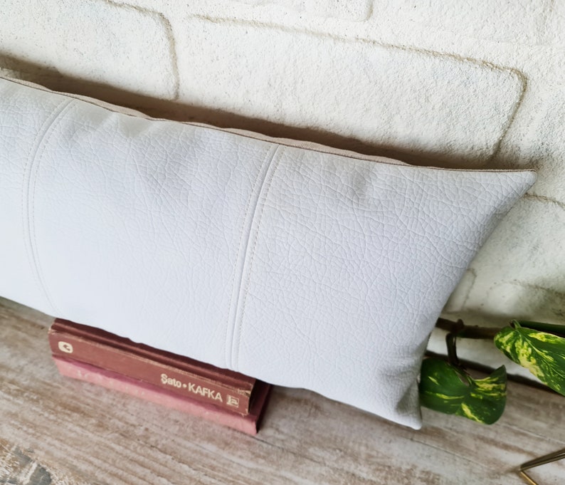 May include: White faux leather lumbar pillow with a subtle textured surface and stitched seams. The pillow is resting on a wooden surface with a book titled "Sato + Kafka" visible.